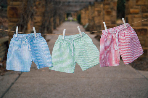 Boys swim trunks in blue seersucker, green seersucker, and red seersucker hanging on a clothing line. 